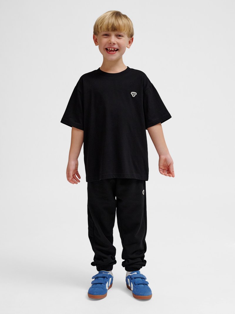 Młode dziecko stoi uśmiechając się, nosi czarny oversize t-shirt i spodnie z niebieskimi sneakersami, na jednolitym jasnym tle.