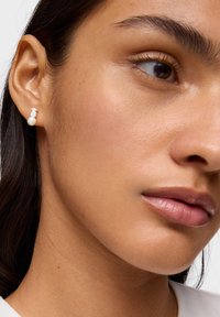 Zbliżenie na twarz kobiety, pokazujące srebrny kolczyk w kształcie małego misia z białą perłową zawieszką.