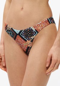 Kobieta w kolorowym, kwiatowo-geometrycznym wzorze na dolnej części bikini, pokazana od talii do połowy uda na białym tle.