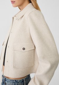 Frau in beiger, taillierter Jacke mit Druckknöpfen und Klappentaschen, kombiniert mit hellblauen Jeans, seitlich über der Hüfte gezeigt.