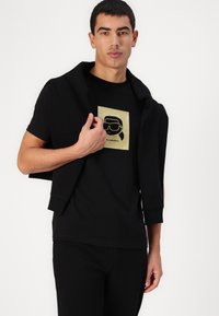 Schwarzes Baumwoll-T-Shirt mit einem goldenen Grafikdesign, das ein stilisiertes Gesicht mit Sonnenbrille zeigt. Das Model hält einen schwarzen Pullover lässig über die Schultern.