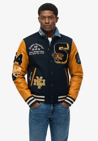 Chaqueta varsity azul marino y naranja con detalles bordados, que presenta puños a rayas, botones de presión y acentos gráficos en las mangas.