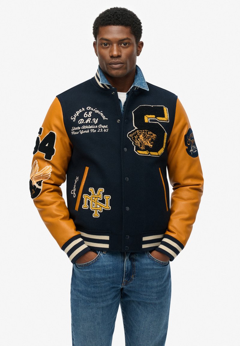 Chaqueta varsity azul marino y naranja con detalles bordados, que presenta puños a rayas, botones de presión y acentos gráficos en las mangas.