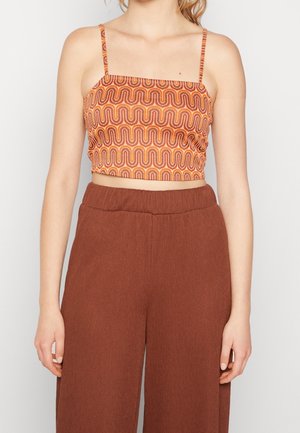 Vrouw draagt een oranje patroon crop top met spaghettibandjes en hoog getailleerde losse bruine broek tegen een effen achtergrond.