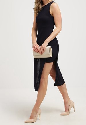 Kobieta w czarnej, asymetrycznej sukience bez rękawów, trzymająca beżową kopertówkę, ubrana w beżowe szpilki, idzie na jasnym tle.
