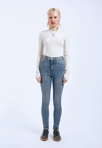 Top blanco de manga larga ajustado combinado con jeans azul de tiro alto y zapatillas gris, con acentos verdes y una textura suave.