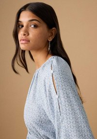 Junge Frau mit langen dunklen Haaren und Sommersprossen, die ein hellblau gemustertes Oberteil mit geknöpften Ärmelschlitzen und hängenden Ohrringen trägt.