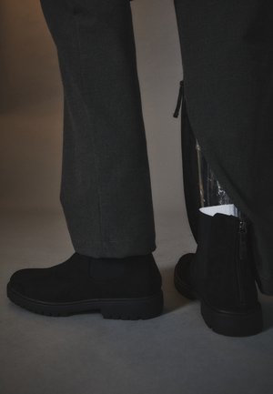 Bottes basses en daim noir portées avec un pantalon sombre, une jambe de pantalon partiellement dézippée révélant une doublure plastique à l'intérieur de la botte.