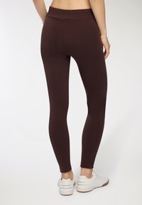 Brązowe legginsy wykonane z elastycznego materiału, z wysokim stanem i gładką fakturą. Połączone z białymi butami sportowymi.