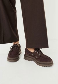 Tmavě hnědé sukně s rounded toe, robustní gumovou podešví a kovovými očky pro tkaničky, spárované s širokými hnědými kalhotami.