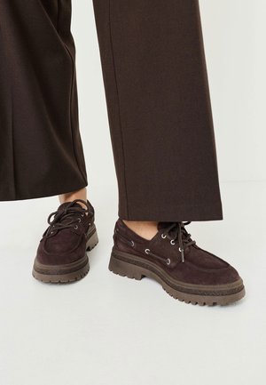 Dunkelbraune Wildlederschuhe mit runder Zehenpartie, robuster Gummisohle und Metallösen für die Schnürsenkel, kombiniert mit weiten braunen Hosen.