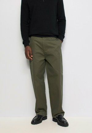 Hombre con pantalones verde oliva, suéter negro y zapatos negros, de pie con una mano en el bolsillo contra un fondo blanco liso.