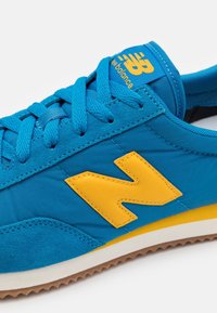 Baskets en textile et daim bleu avec un logo ‘N’ en caoutchouc jaune, lacets plats et une semelle intermédiaire blanche avec une semelle extérieure en caoutchouc.