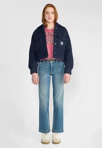 Veste courte bleu marine en tissu texturé, avec deux poches avant, des boutons noirs et un logo en forme de cœur. Portée sur un t-shirt graphique rose et un jean bleu.