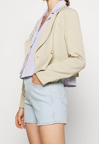 Žena v béžovém krátkém saku, světle fialové blůze a světle modrých džínových šortkách, s jednou rukou v kapse, pózující na bílém pozadí.