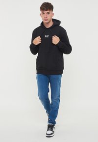 Svart hettegenser med frontlomme og EA7-logo på brystet, parret med blå jeans og svarte joggesko med hvite detaljer.