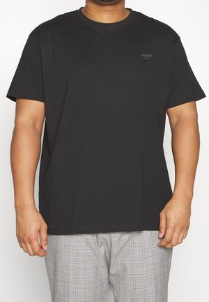 Mann trägt ein schlichtes schwarzes T-Shirt mit kurzen Ärmeln und dem "JOOP! JEANS"-Logo auf der linken Brust sowie graue karierte Hose, steht vor weißem Hintergrund.