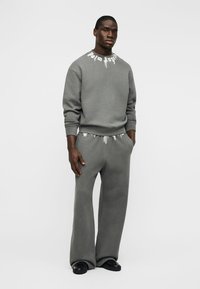 Άνδρας που φοράει γκρι φούτερ και παντελόνια φαρδιά με λευκές λεπτομέρειες, στέκεται με τα χέρια στις τσέπες μπροστά σε απλό φόντο.