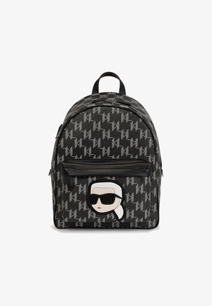 Mochila negra con patrón repetitivo blanco KL y bolsillo frontal que presenta una cara de dibujo animado con cabello blanco y grandes gafas de sol negras.