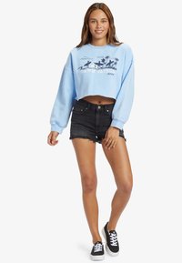 Sweatshirt corto azzurro con grafica "DAWN PATROL", abbinato a pantaloni denim neri e sneakers nere con lacci bianchi.