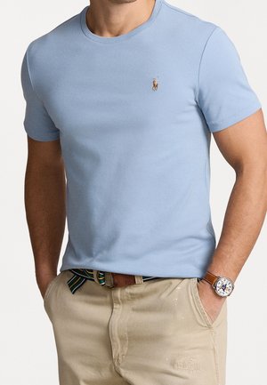 Mann trägt ein hellblaues T-Shirt mit kurzen Ärmeln und kleinem Logo eines Pferdes mit Reiter, beige Hosen, gestreiften Gürtel und Armbanduhr mit braunem Lederarmband.