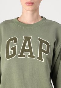 Olivově zelený svetr s žebrovaným kulatým výstřihem. Zdůrazněno výrazným nápisem "GAP" v tmavší barvě, orámovaným bílým prošíváním. Měkká textura.