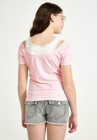 Lyserød ribbet kortærmet top med åben skulderdesign, lagd over en hvid tanktop med blondekant, paired med grå denimshorts.