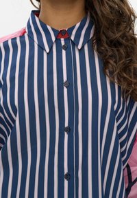Chemise rayée dans des tons navy et rose, avec un col à boutons, des fermetures à boutons noires et des accents de manches contrastants. Texture de tissu lisse.