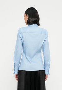 Lys blå skjorte med lange ermer, tilpasset design, knappede mansjetter og sømdetaljer på baksiden. Brukes sammen med et svart plissert skjørt.