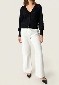 Žena v černém zapínaném pleteném kardiganu, bílých texturovaných širokých kalhotách a černých botách se špičatou špičkou stojí před jednobarevným pozadím.