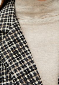 Blazer a quadri su misura in nero, crema e marrone, abbinato a un dolcevita a coste color beige chiaro. Focus sul contrasto dei materiali e dei motivi.