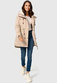 Beige parka jas met een capuchon, bontvoering, trekkoordsluiting en houten accent toggels. Gepaard met blauwe jeans en witte sneakers.
