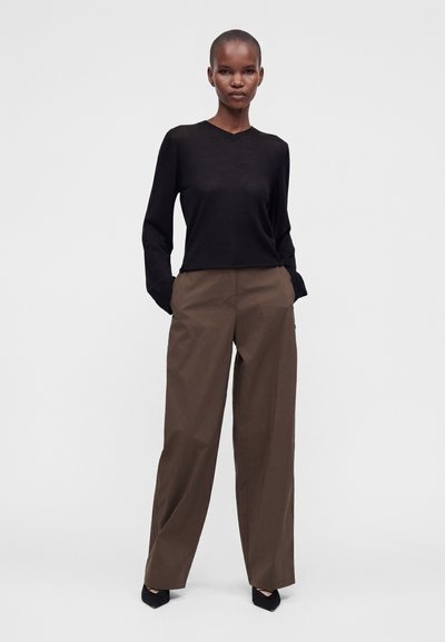Femme debout les mains dans les poches, portant un haut noir à manches longues, un pantalon marron à jambes larges et des chaussures noires pointues.