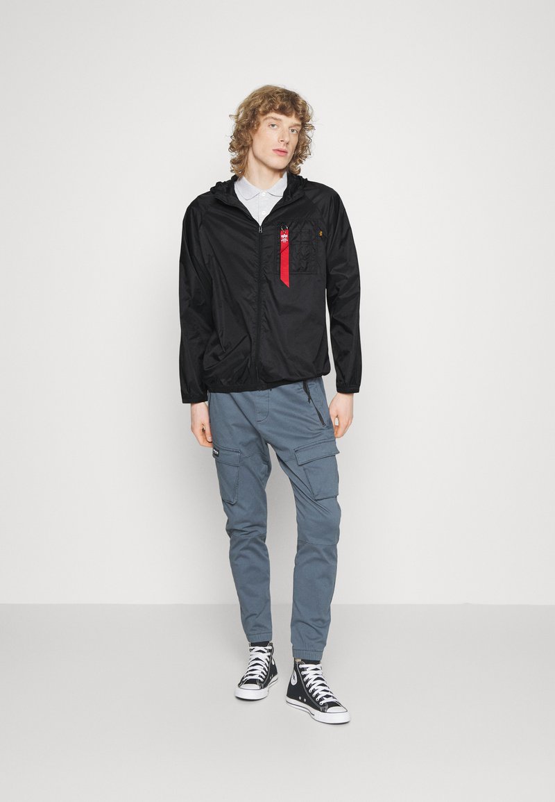 Schwarze Zip-Jacke mit Kapuze, rote Bänderdetails auf der Brusttasche, kombiniert mit grauen Cargo-Hosen und schwarzen High-Top-Sneakern.