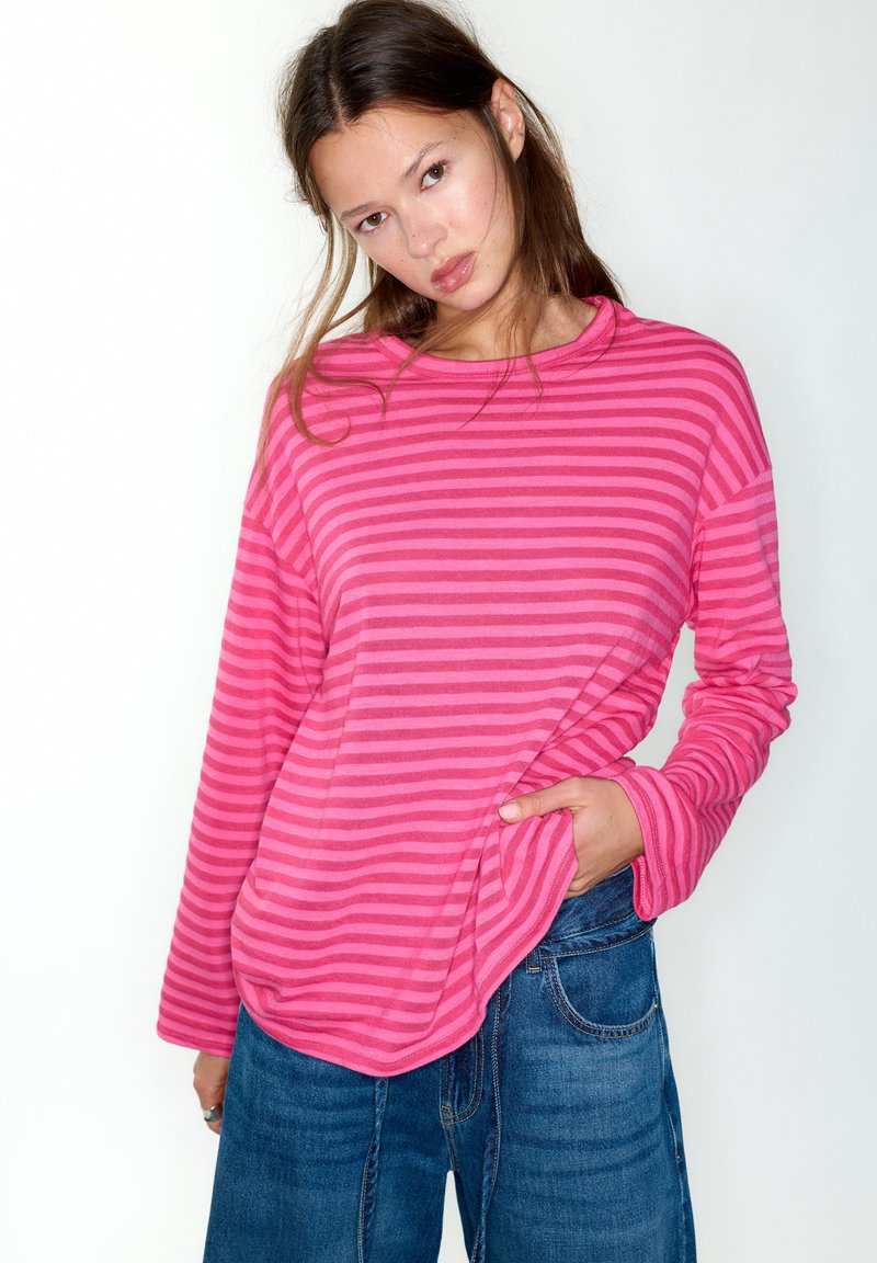 Pink langærmet top med striber, afslappet pasform og rund halsudskæring, kombineret med blå jeans. Blødt stof og synlige syningsdetaljer.