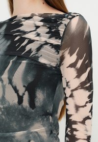 Gros plan d'une personne portant un haut transparent à manches longues noir et beige avec un motif tie-dye, épaule et haut du bras visibles.