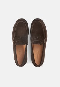 Καφέ μοκασίνια από δέρμα σουέτ με χαμηλό προφίλ, ραφές λεπτομέρειας και διακοσμητικό λουράκι κατά μήκος της πάνω πλευράς. Λείος εσωτερικός επένδυση.