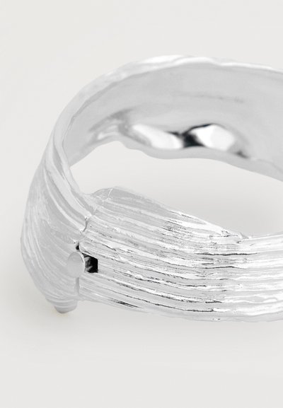 Bracelet manchette texturé argent avec un design rainuré et une petite fermeture ronde sur fond blanc.