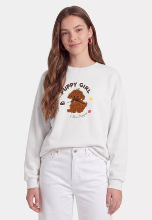 Ung kvinde med langt brunt hår bærer pink øreringe i ring og hvid sweatshirt med en brun hvalp og tekst "Puppy Girl I Love Puppies."