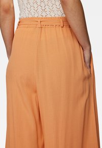 Oranje plooibroek van een gestructureerde stof, met zijzakken en een ceintuur in de taille. Het ontwerp is vloeiend en lichtgewicht.