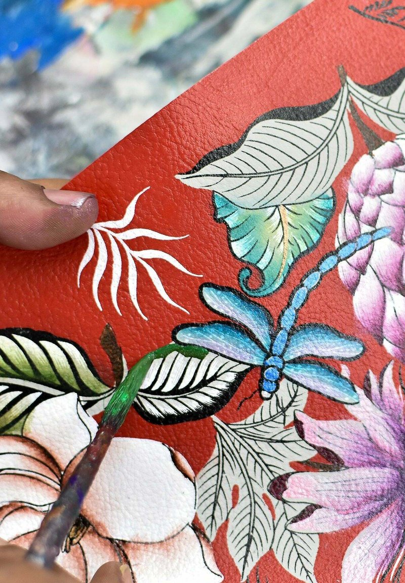 Lederoberfläche mit lebhaft rotem Hintergrund, der mit detaillierten Blumen- und Blattmustern verziert ist. Eine blaues Libelle ist prominent dargestellt, mit einem Pinsel in der Nähe.