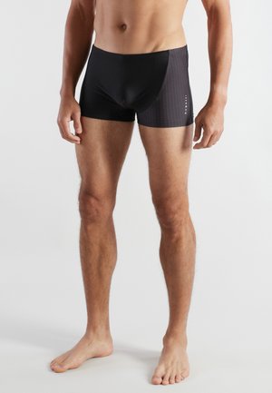 Male torso and legs wearing black and dark gray fitted swim trunks, standing barefoot against a plain light background.