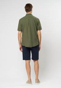 Camicia verde oliva a maniche corte, tessuto leggero, dettaglio della piega sul retro. Abbinata a pantaloni corti blu navy, calzature neutre, design minimale.