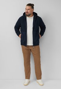 Mężczyzna w granatowej, rozpinanej bluzie z kapturem, białej koszulce, brązowych spodniach i białych trampkach, stojący z rękami w kieszeniach na tle gładkiej ściany.