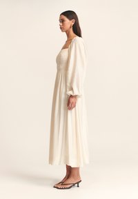 Cremefarbendes langes Kleid mit einem taillierten, gerafften Oberteil und Puffärmeln. Der fließende Rock reicht bis zur Mitte der Wade. Kombiniert mit schwarzen Sandalen mit Absatz.