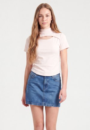 Junge Frau mit langen Haaren, die ein hellrosa kurzärmeliges Oberteil mit Ausschnittdetail und einen blauen Jeansrock trägt, stehend vor einem schlichten hellen Hintergrund.