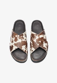 Sandales à glisser en cuir de vachette marron et crème avec des sangles croisées, présentant une semelle noire plate et une semelle intérieure en cuir lisse. Aucune quincaillerie ni accents visibles.