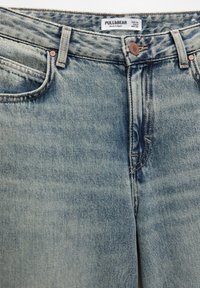 Gros plan sur un jean en denim bleu clair avec un bouton avant visible, des passants de ceinture, des poches avant et une étiquette de marque indiquant la taille et l'origine.