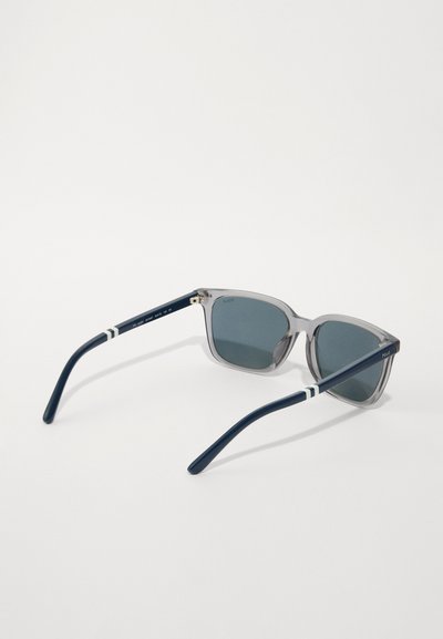 Ochelari de soare transparenți gri cu lentile închise, având brațe bleu marin cu dungi albe. Design cu ramă pătrată și accente metalice.
