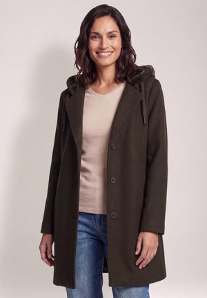 Mujer sonriendo, vestida con un abrigo marrón oscuro con capucha forrada de piel, camiseta beige y vaqueros azules sobre un fondo simple.